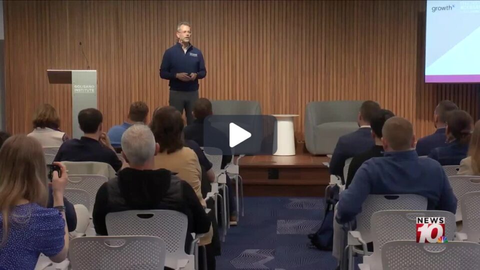 A man addresses a crowd in a business-style presentation setting. 