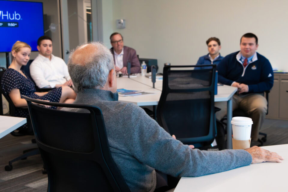 An instructor speaks to a group of college students and faculty around a classroom table. 