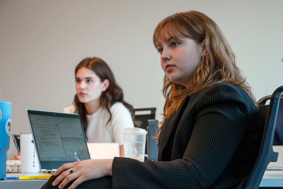 Two female students listen to an instructor with a laptop on shared table. 