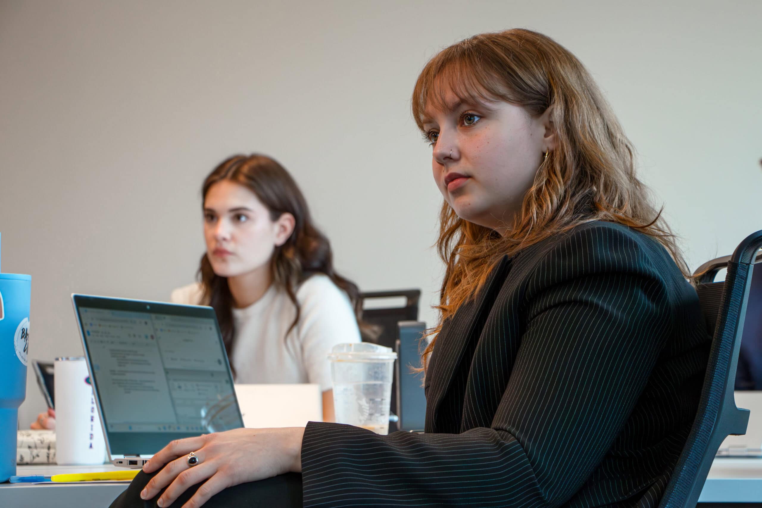 Two female students listen to an instructor with a laptop on shared table.