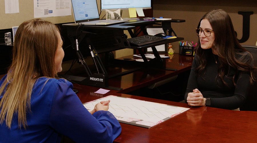 Two woman sitting at a desk in an office having a conversation.