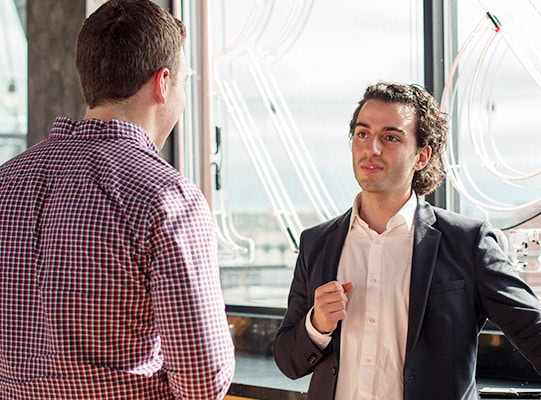 Two men speaking in front of a large window on a sunny day.