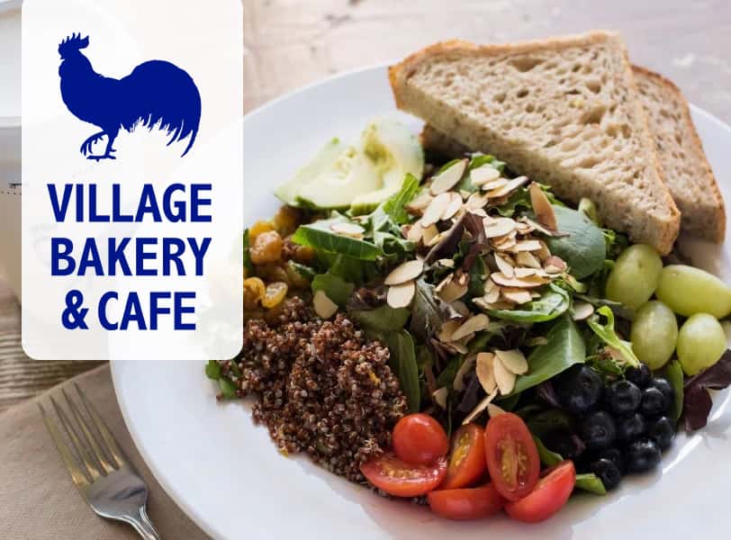 Fruits, vegetables, and sandwich on a plate with the Village Bakery & Cafe logo.