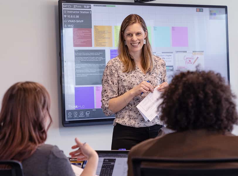 A teacher stands in front of a digital screen while speaking with two students.
