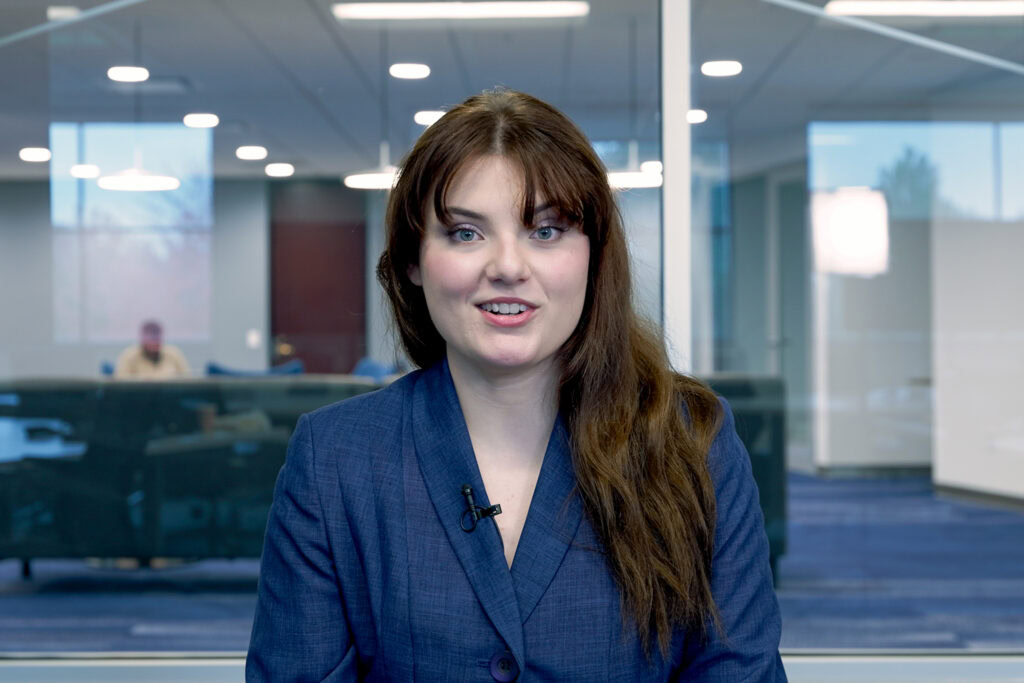 A female student looks straight at the camera in a modern classroom environment.