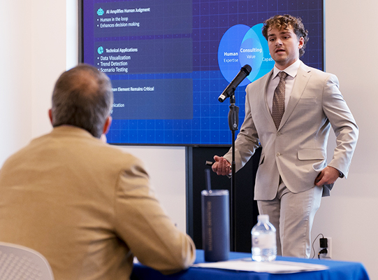 A man in a suit stands at a microphone in front of a screen delivering a presentation.