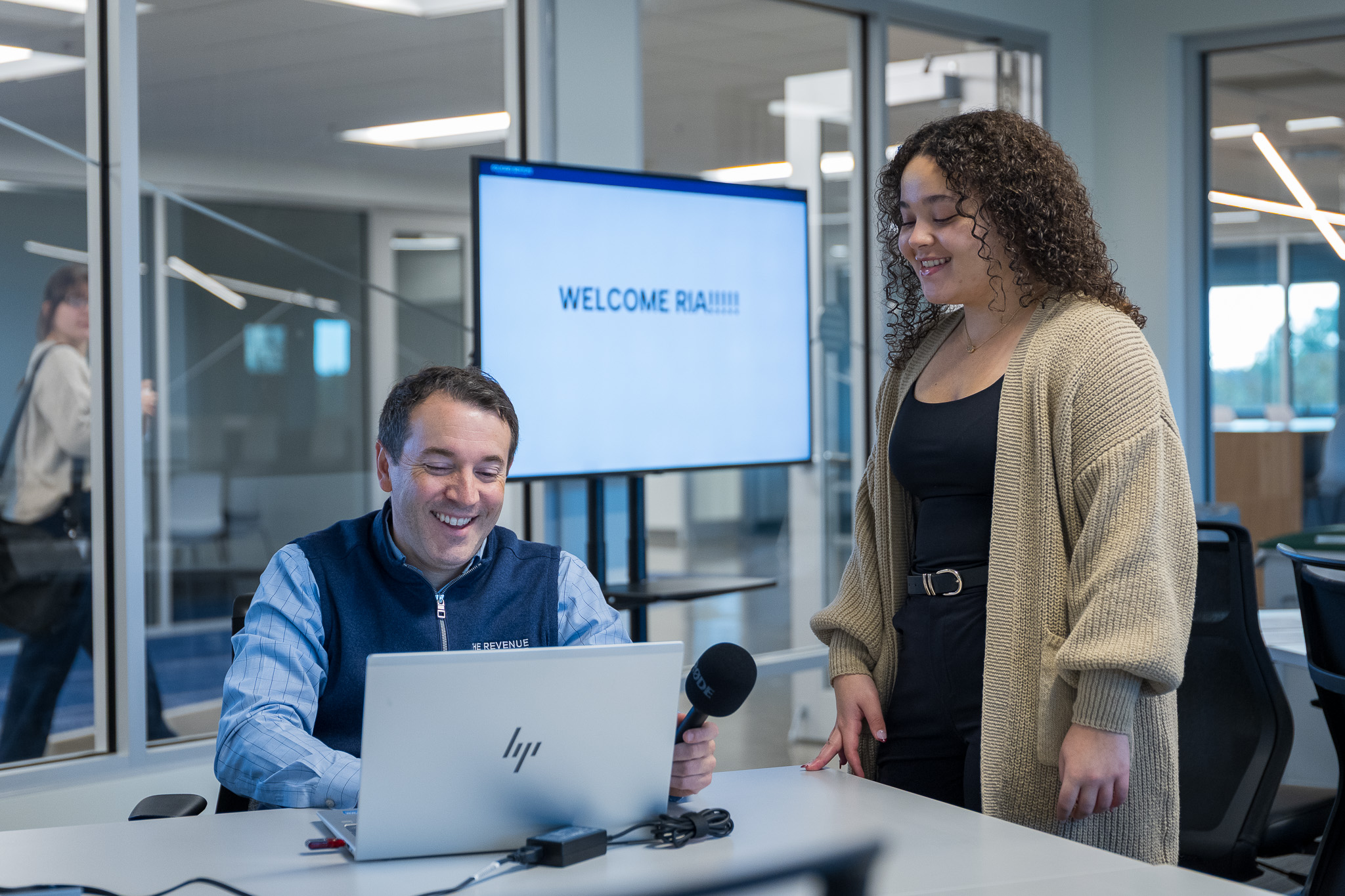 A man seated at a table smiling while looking at a laptop, holding a handheld microphone, as a woman stands beside him smiling. They are in a modern office or classroom with glass walls, and a screen in the background displays a welcome message.