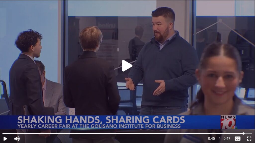 People networking at an indoor career fair, with individuals conversing and gesturing in a professional setting. A television news lower-third reads ‘Shaking Hands, Sharing Cards’ and ‘Yearly Career Fair at the Golisano Institute for Business,’ with a local news logo.