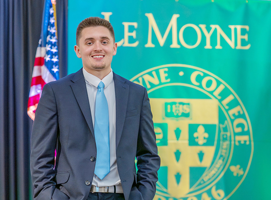Nicholas Figler poses for a photo in front of an American flag and a LeMoyne college.