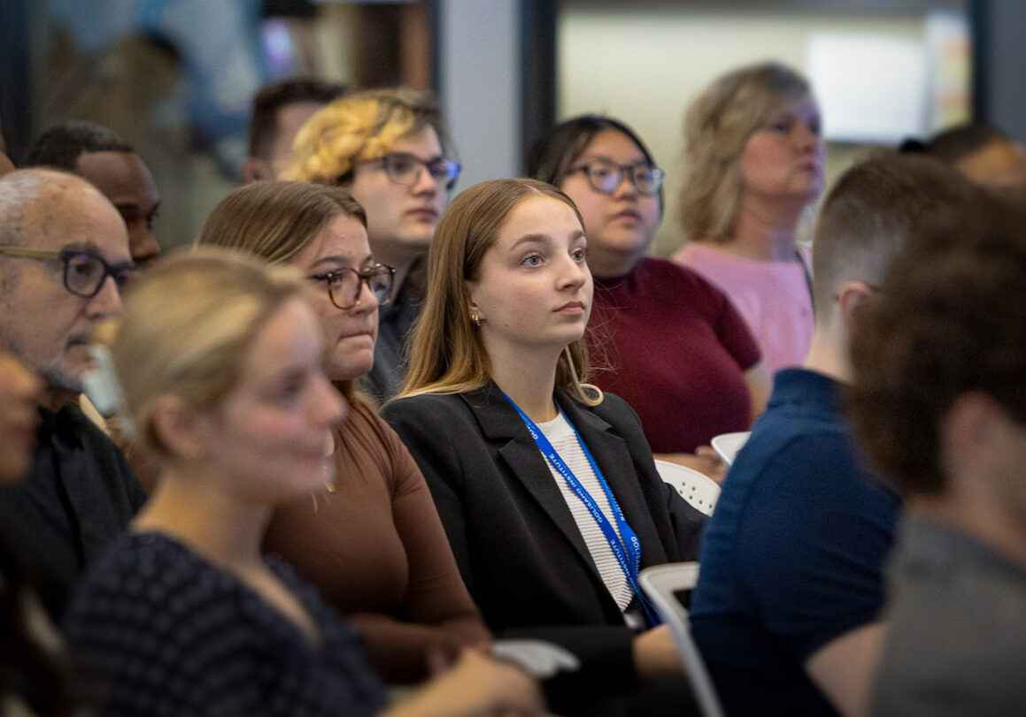 A group of people sit and listen to a speaker off camera